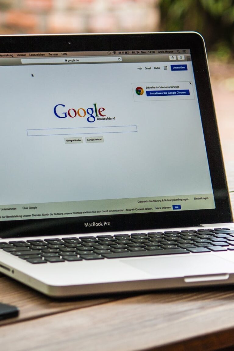 A MacBook Pro displaying Google Search on a wooden table outdoors, next to a smartphone.